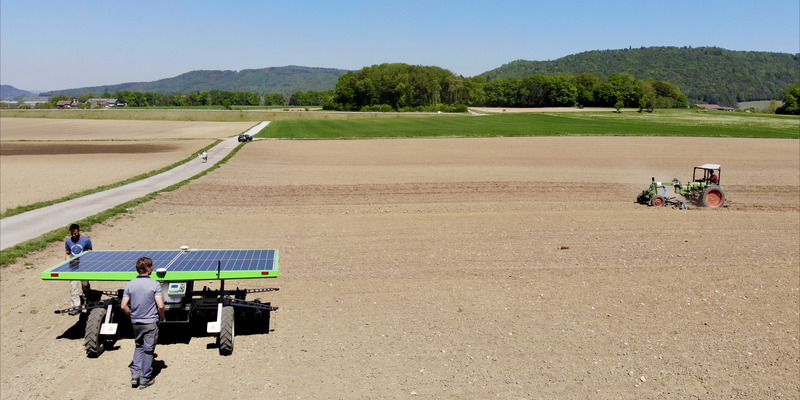 Zwei junge Männer gehen mit einer Maschine über einen trockenen Acker mit Traktor im Hintergrund.